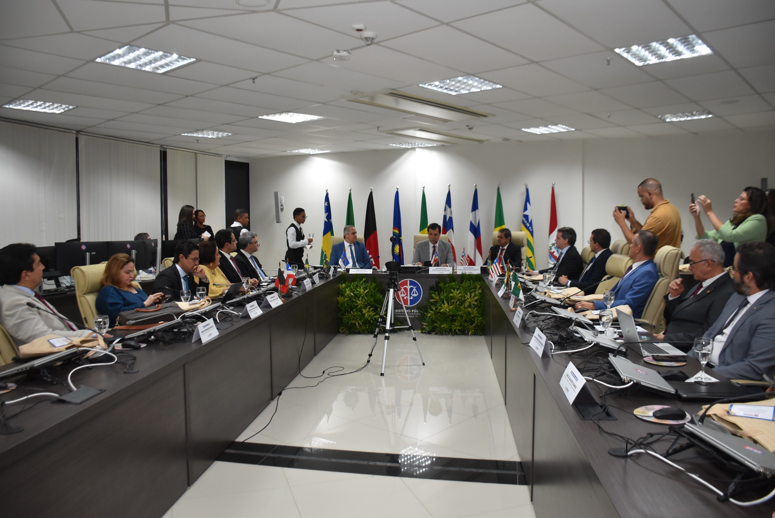 Procuradores-gerais da região Nordeste e outras autoridades reunidos sentado a uma mesa marrom escura, com bandeiras dos estados ao fundo contrastando com parede branca.